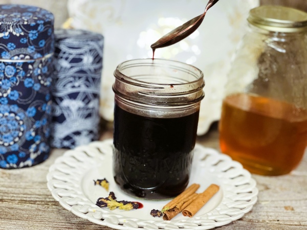 Elderberry syrup dropping off of a spoon into a mason jar, sitting on a white ceramic plate topped with dried flowers and cinnamon sticks.